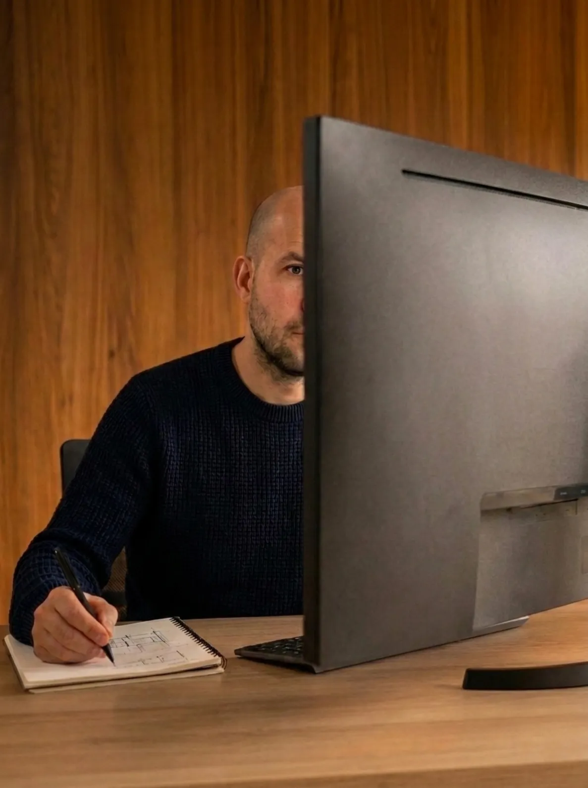 A drafter sketching a floor plan by hand in a notebook beside a large monitor, in a wood-panelled studio.